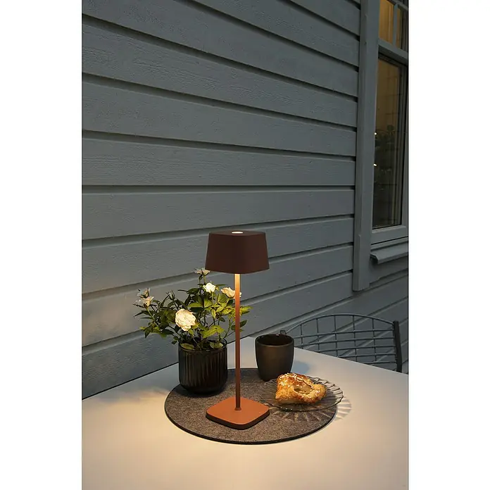 Konstsmide Akku-Tischleuchte Capri - 2,2 W, L x B x H: 10 x 10 x 36 cm, Braun - Ein stimmungsvolles Bild einer Tischlampe, die eine gemütliche Atmosphäre auf einem Tisch mit Gebäck und Getränk erzeugt.