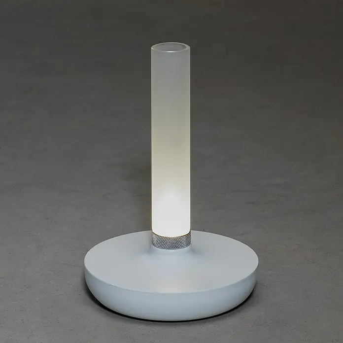 Konstsmide Akku-Tischleuchte Biarritz - 2,5 W, L x B x H: 13,5 x 13,5 x 20,5 cm, Weiß, Warmweiß - Nahaufnahme einer modernen weißen Tischleuchte mit zylindrischem Lampenschirm und silberfarbenem Akzent auf einem grauen Untergrund.