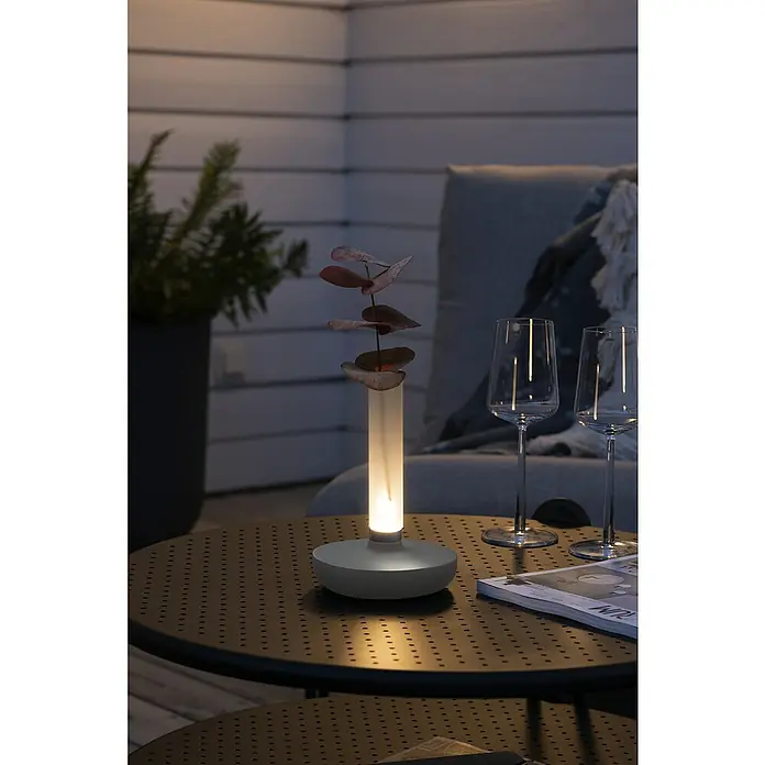 Konstsmide Akku-Tischleuchte Biarritz - 2,5 W, L x B x H: 13,5 x 13,5 x 20,5 cm, Weiß, Warmweiß - Eine moderne Tischleuchte mit grauem Sockel und einem transparenten Aufsatz, in dem eine einzelne Pflanze steht. Die Lampe strahlt warmes Licht aus.
