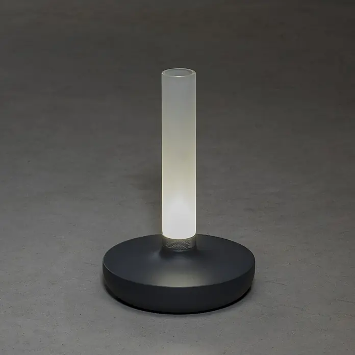 Konstsmide Akku-Tischleuchte Biarritz - 2,5 W, L x B x H: 13,5 x 13,5 x 20,5 cm, Grau, Warmweiß - Ein minimalistisches Bild zeigt eine moderne Tischleuchte mit einem zylindrischen Schirm auf einem runden Sockel, die ein sanftes Licht verbreitet.