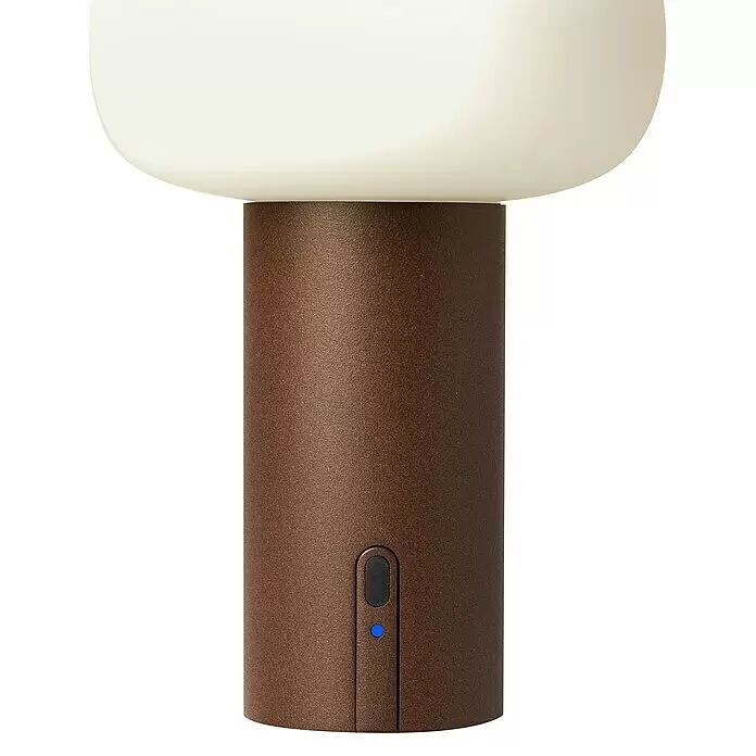 Konstsmide Akku-Tischleuchte Antibes - 2,5 W, L x B x H: 13 x 13 x 19 cm, Rost, Mehrfarbig - Eine tragbare Lampe mit cremefarbener Oberseite und einem strukturierten, dunkelrotbraunen zylindrischen Sockel mit schwarzem Knopf und blauer Anzeigeleuchte.