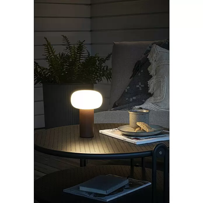 Konstsmide Akku-Tischleuchte Antibes - 2,5 W, L x B x H: 13 x 13 x 19 cm, Rost, Mehrfarbig - Dunkelbraune portable Lampe mit ovalem, leuchtendem weißem Diffusor auf perforiertem Tisch, beleuchtet einen Außenbereich.