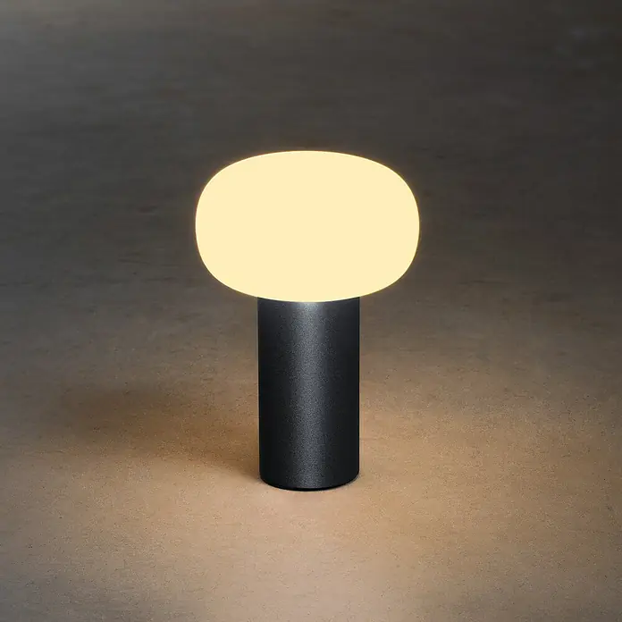 Konstsmide Akku-Tischleuchte Antibes - 2,5 W, L x B x H: 13 x 13 x 19 cm, Schwarz, Mehrfarbig - Nahaufnahme einer modernen Tischleuchte mit einem runden Lampenschirm auf einem zylindrischen Sockel, die ein warmes Licht ausstrahlt.