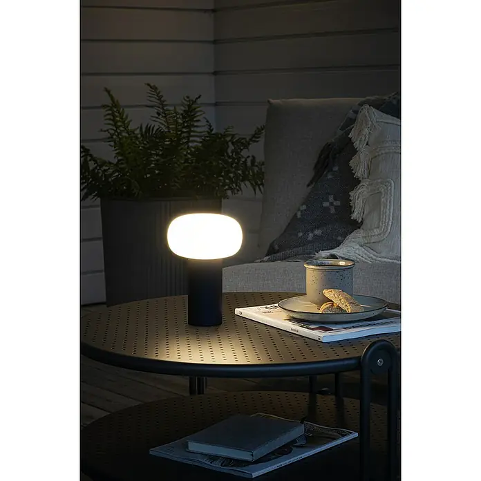 Konstsmide Akku-Tischleuchte Antibes - 2,5 W, L x B x H: 13 x 13 x 19 cm, Schwarz, Mehrfarbig - Stimmungsvolle Tischleuchte auf einem Gartentisch mit Kaffeetasse und Zeitschriften im Hintergrund.