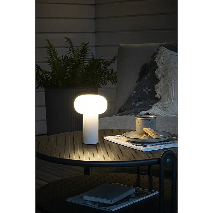 Konstsmide Akku-Tischleuchte Antibes - 2,5 W, L x B x H: 13 x 13 x 19 cm, Weiß, Mehrfarbig - Nahaufnahme einer stilvollen, kabellosen Lampe auf einem Tisch mit Getränken und Zeitschriften im Freien, perfekt für eine entspannte Atmosphäre.