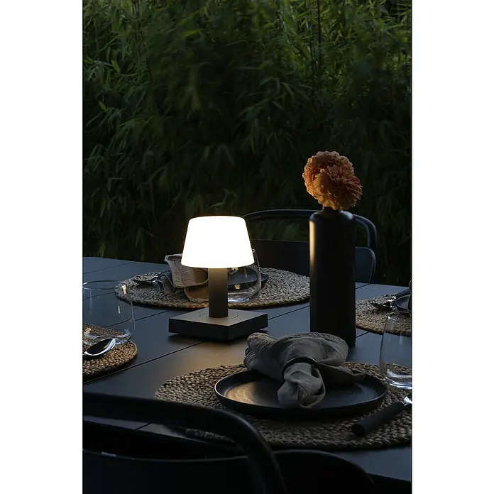 Konstsmide Akku-Tischleuchte Monaco - 2,5 W, L x B x H: 12,5 x 12,5 x 19 cm, Grau, Warmweiß - Elegante Tischdekoration mit einer modernen Lampe, einem schwarzen Teller mit Serviette und einer Vase mit Blume, die eine warme Atmosphäre schafft.