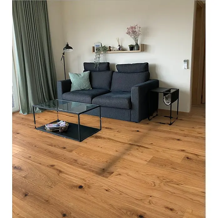 Parkett - Eiche Yankee, Landhausdiele, 1.900 x 190 x 14 mm - Wohnzimmeransicht mit einem stilvollen grauen Sofa, einem Couchtisch mit Glasplatte und einem schwarzen Beistelltisch neben dem Sofa.