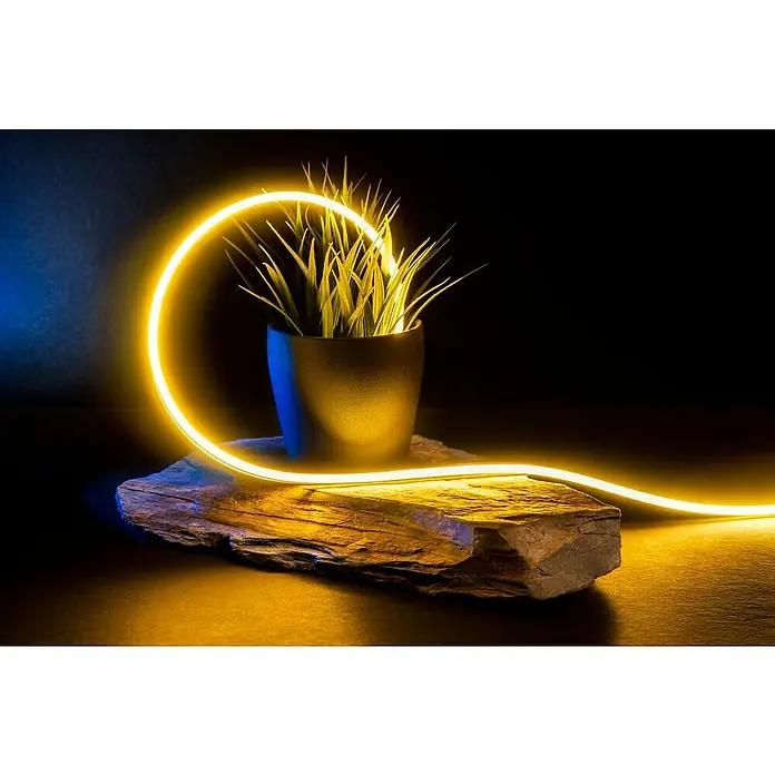 Alverlamp Tira LED a metros Neon - Largo: 100 m, Color de luz: Blanco cálido, 9 W, 1.308 lm - Primer plano de una planta artificial en una maceta, rodeada por un tubo de neón amarillo brillante sobre una roca, mostrando una pieza de decoración moderna.