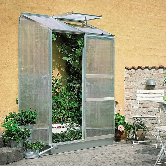 Halls Gewächshaus Altan 2 - Außenmaß inkl. Dachüberstand (B x T): 131 x 68 cm, Material Seitenwand: Kunststoff, 4 mm, Silbergrau - Nahaufnahme eines Anlehngewächshauses mit geöffneter Tür, gefüllt mit üppigen Pflanzen, das an einer gelben Backsteinmauer steht.