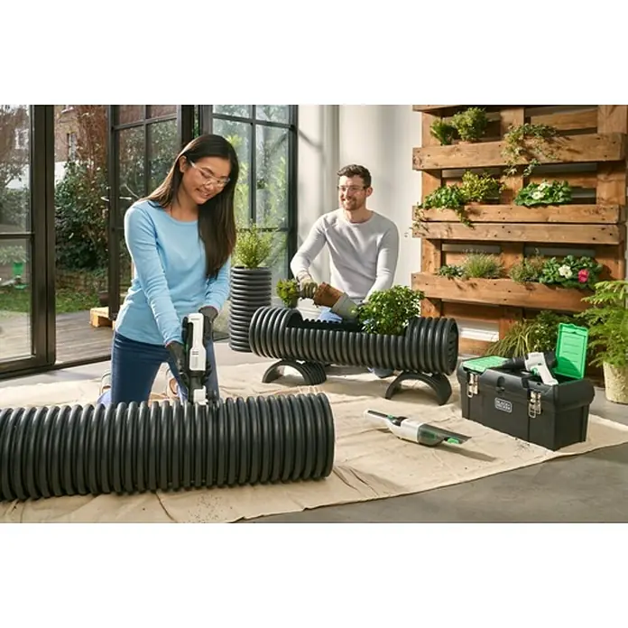 Black+Decker reviva Caja para herramientas - 19'', Polipropileno - Dos personas crean jardineras creativas con tubos de plástico negros, con una caja de herramientas y una aspiradora de mano cerca.