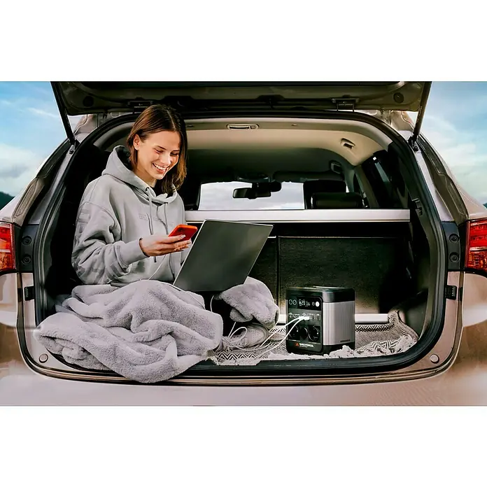 Yard Force Powerstation LX PS600 - 518 Wh, Nennleistung: 600 W, Max. Leistung: 1.200 W - Eine junge Frau sitzt im Kofferraum eines Autos, mit einem Laptop und einem Handy, während eine RapidForce Powerstation im Hintergrund zu sehen ist.