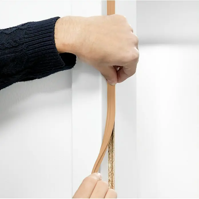 Primo Türdichtung Lignum 34 - Beige, 5 m, Passend für: Nutbreite 3 – 4 mm - Eine Person bringt eine braune Kantenumleimer an einer weißen Oberfläche an. Perfekt zum Schutz und zur ästhetischen Aufwertung von Möbeln.