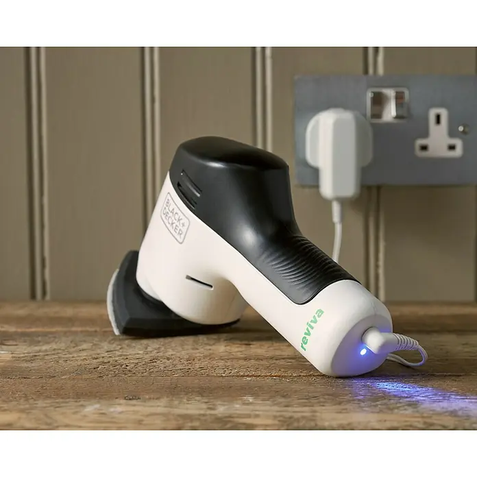 Black+Decker reviva Lijadora delta de batería - 12 V, 1,5 Ah, 11.000 r.p.m. - Primer plano de una lijadora inalámbrica Black+Decker Reviva sobre una mesa de madera, enchufada a una toma de corriente. El dispositivo es blanco con detalles negros.
