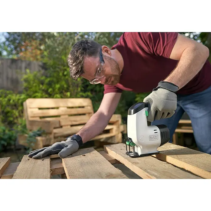 Black+Decker reviva Sierra de calar de batería - 12 V, 1 batería, 1,5 Ah - Un hombre corta madera con una sierra de calar Black+Decker, lleva guantes y gafas de seguridad. Al fondo hay una pila de madera.