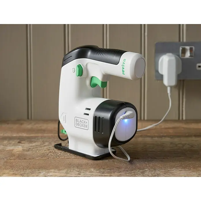 Black+Decker reviva Sierra de calar de batería - 12 V, 1 batería, 1,5 Ah - Primer plano de una sierra de calar BLACK+DECKER Reviva sentada en una mesa de madera y enchufada a una toma de corriente para cargar.