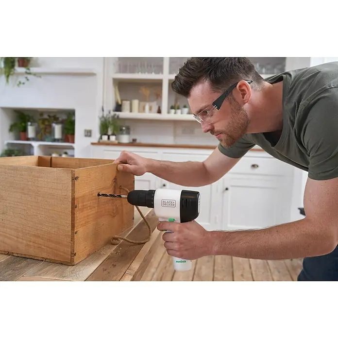 Black+Decker reviva Taladro atornillador de batería - 12 V, 1 batería, 1,5 Ah, 26 Nm - Un hombre perfora una caja de madera con un taladro inalámbrico Black+Decker Reviva. ¡Gafas de seguridad para protección!