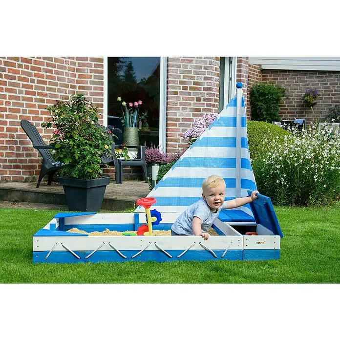 Dobar Sandkasten Maritim - 140 x 120 x 117 cm, Holz, Blau/Weiß - Ein entzückender Junge spielt in einem blauen und weißen Dobar-Sandkasten mit Segel, umgeben von üppigem Grün. Perfekt für Gartenspaß!