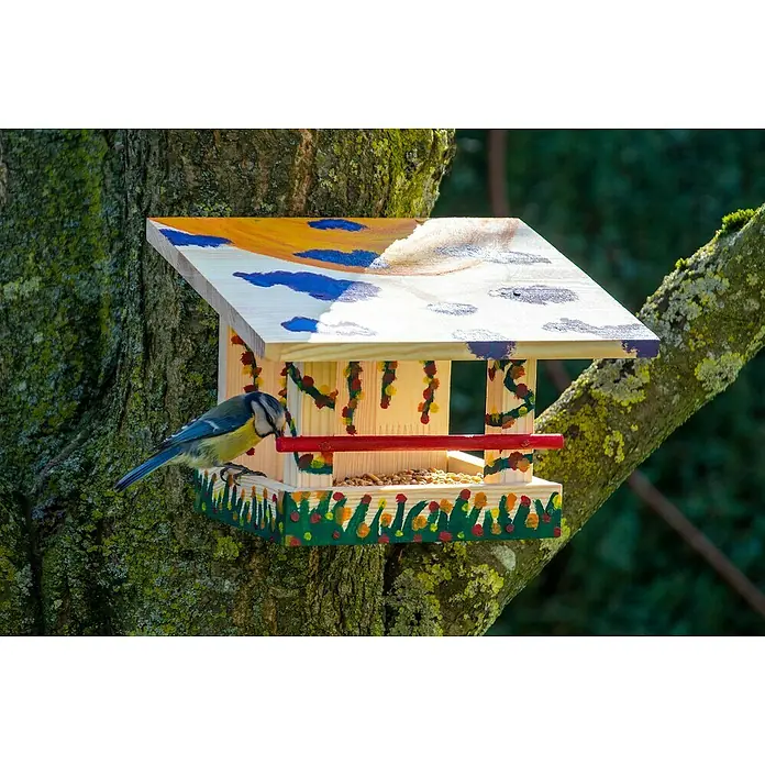 Dobar Vogelhaus Bausatz - 22,5 x 18 x 16 cm, Holz, Natur - Eine Blaumeise sitzt auf einem bunten Vogelhaus aus Holz, das mit fröhlichen Motiven bemalt ist und im Garten an einem Baum hängt.