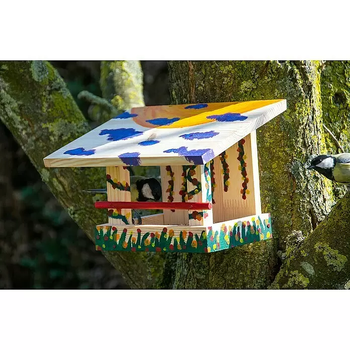 Dobar Vogelhaus Bausatz - 22,5 x 18 x 16 cm, Holz, Natur - Ein bunt bemaltes Vogelhäuschen aus Holz mit Sonne, Wolken und Blattwerk hängt an einem Baumstamm. Zwei Meisen nutzen es.