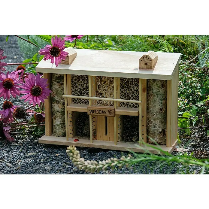 Dobar Insektenhotel Alabama44 x 13,5 x 30 cm, Kiefer, Natur - Ein helles Holzinsektenhotel, umgeben von lila Blüten, bietet verschiedenen Insekten Unterschlupf und Nistmöglichkeiten im Garten.