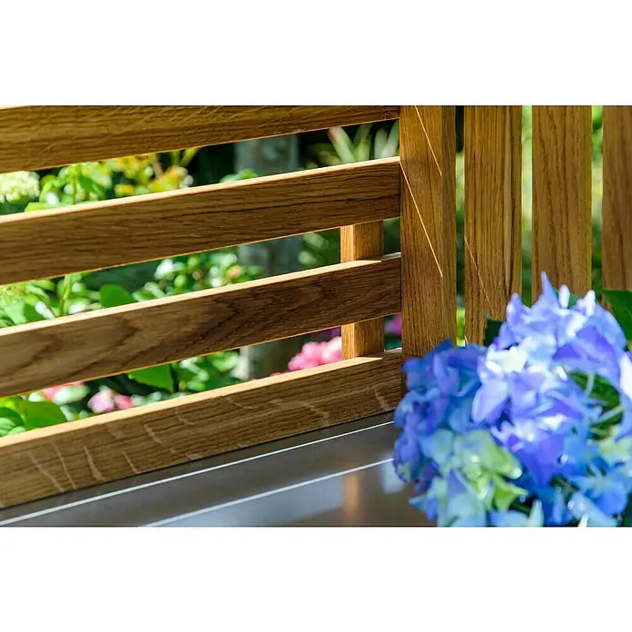 Dobar Pflanztisch Eiche - L x B x H: 117 x 62 x 124 cm - Nahaufnahme eines modernen Holzzauns mit blauen Hortensien im Vordergrund, der einen Garten mit üppigem Grün umrahmt.