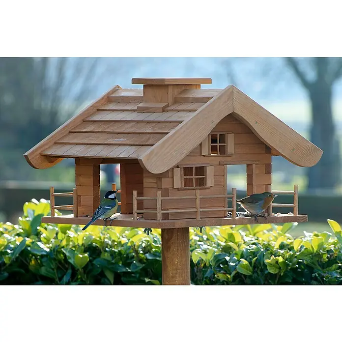 Dobar Vogelfutterhaus Almhütte - L x B x H: 66 x 66 x 136,5 cm, Braun, Holz - Ein hölzernes Vogelhaus mit zwei kleinen Vögeln, die auf der Futterplattform sitzen, umgeben von grünen Büschen.