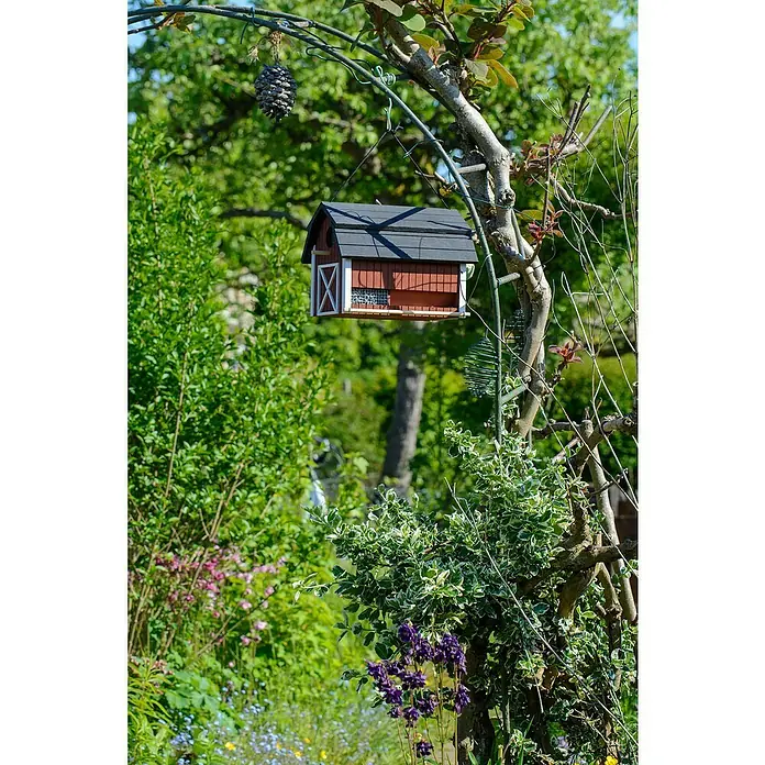 Dobar Vogelhaus Katthult - 31,5 x 21 x 23,5 cm, Holz, Rot - Ein rotes Vogelhaus mit schwarzem Dach hängt an einem Baum, umgeben von Grünpflanzen und Blumen, die einen Gartenblick bieten.