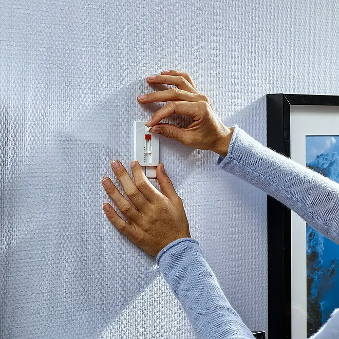Tesa Verstellbarer Klebenagel - Geeignet für: Tapeten, Belastbarkeit: 2 kg, 2 Stk., Weiß - Nahaufnahme einer Person, die einen Powerstrip-Nagel von tesa in eine strukturierte weiße Wand einführt, um Bilder aufzuhängen.