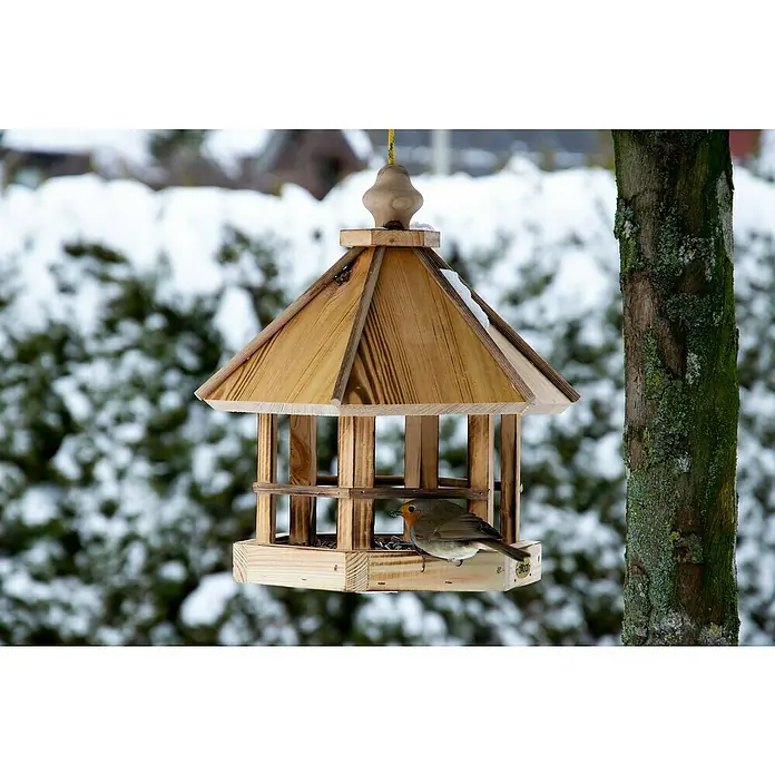 Dobar Vogelhaus Caliente - 29 x 29 x 34 cm, Holz, Braun - Ein hölzernes Vogelhaus mit einem kleinen Vogel darin, der Futter sucht, inmitten einer verschneiten Winterlandschaft.