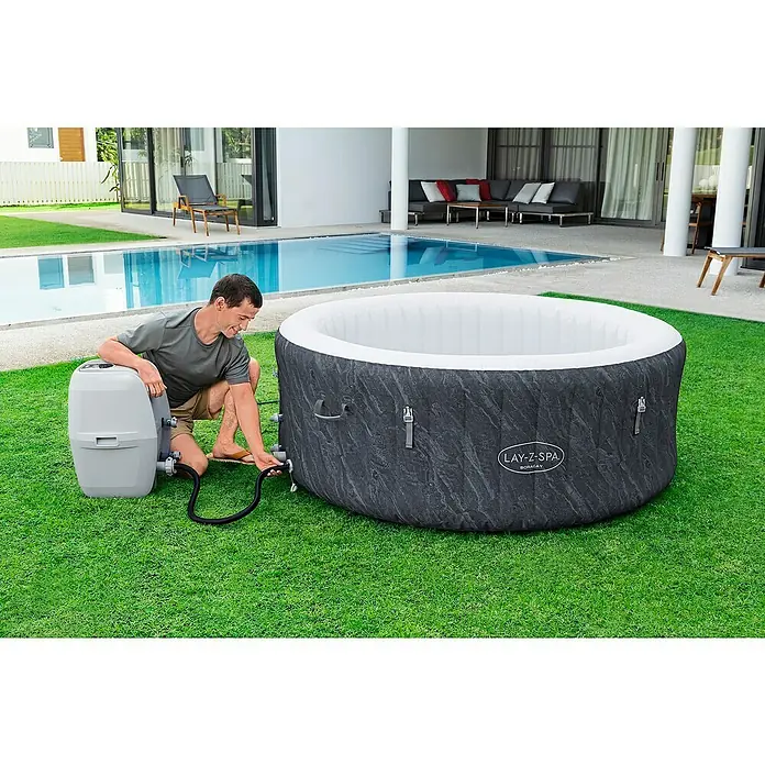 Bestway Lay-Z-Spa Mobil-Whirlpool Boracay Air Jet - Ø x H: 180 x 66 cm, Schwarz, 669 l - Ein Mann schließt einen Lay-Z-Spa Boracay Whirlpool an eine Pumpe an. Der Pool ist auf einem Rasen neben einem Pool aufgestellt.