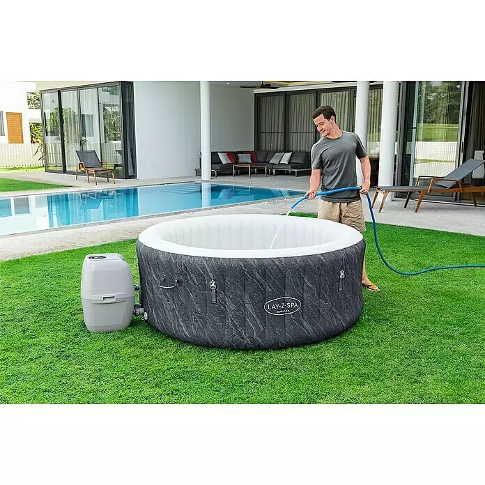 Bestway Lay-Z-Spa Mobil-Whirlpool Boracay Air Jet - Ø x H: 180 x 66 cm, Schwarz, 669 l - Mann füllt einen dunkelgrau marmorierten aufblasbaren Whirlpool mit Wasser. Eine graue Pumpeneinheit ist angeschlossen.