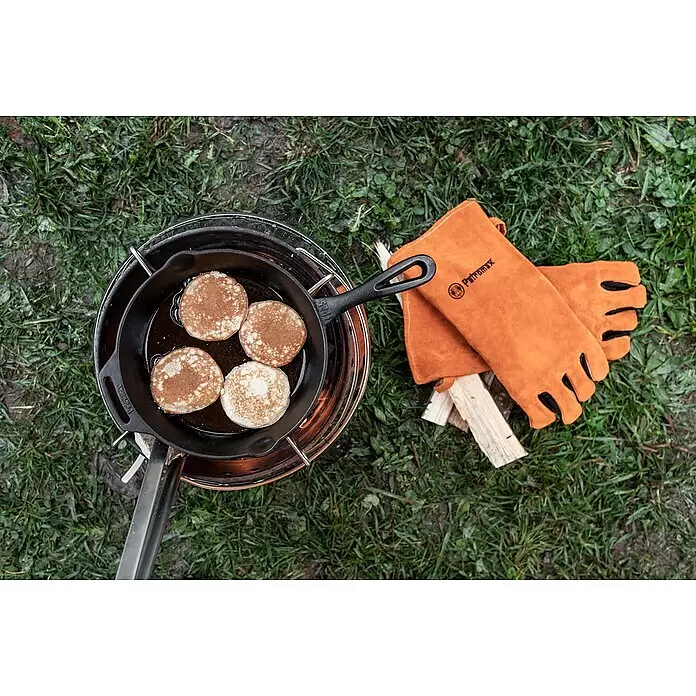 Petromax Grillpfanne - Ø x H: 30 x 4,5 cm, Gusseisen - Gusseisenpfanne brät Pfannkuchen auf einem Campingkocher neben orangefarbenen Hitzeschutzhandschuhen und Anzündholz.