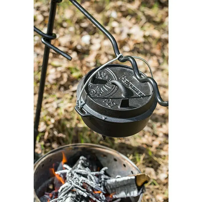 Petromax Dutch Oven - Ø x H: 25,2 x 16 cm, Gusseisen, 1,6 l, Mit Füßen - Ein Petromax Feuertopf hängt über einem Lagerfeuer, bereit für kulinarische Abenteuer im Freien. Ideal für Camping und Outdoor-Küche!