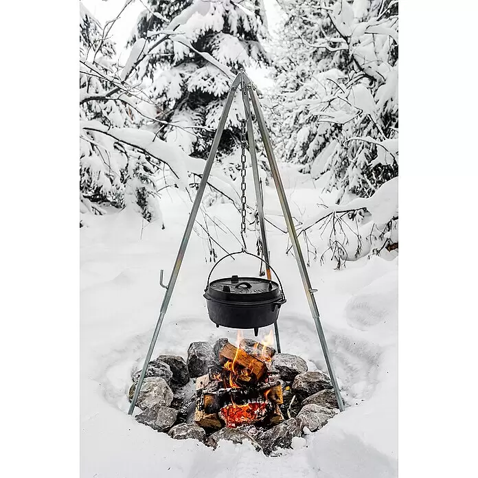 Petromax Dreibeinständer - Grau - Ein silbernes Metall-Kochstativ hält einen schwarzen Gusseisen-Dutch Oven über einem Lagerfeuer im verschneiten Wald.
