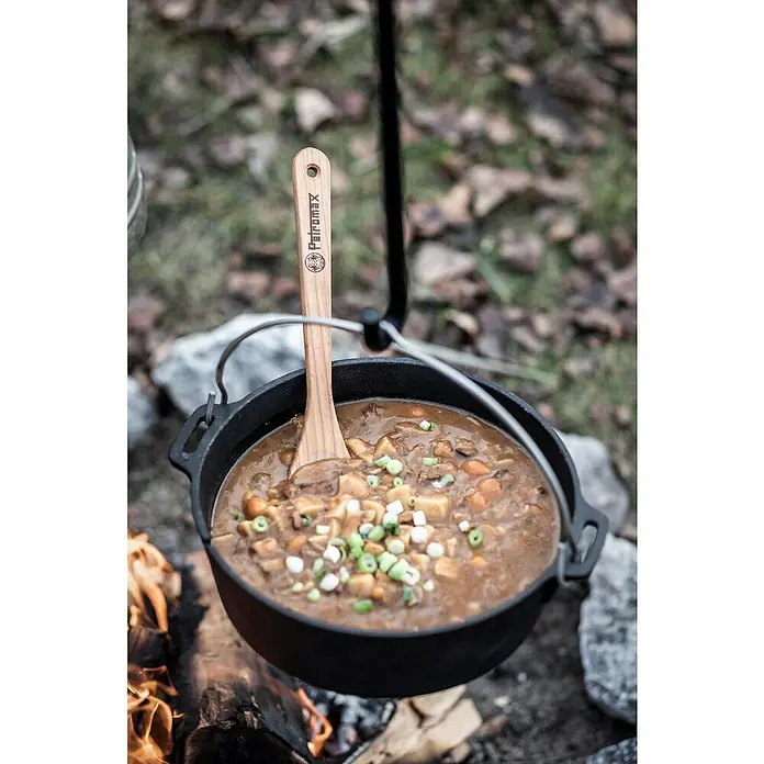 Petromax Dutch Oven - Ø x H: 42 x 24 cm, Gusseisen, 10,8 l, Mit Füßen - Ein Petromax Dutch Oven hängt über einem Lagerfeuer, gefüllt mit Eintopf und einem Holzlöffel der Marke Petromax.