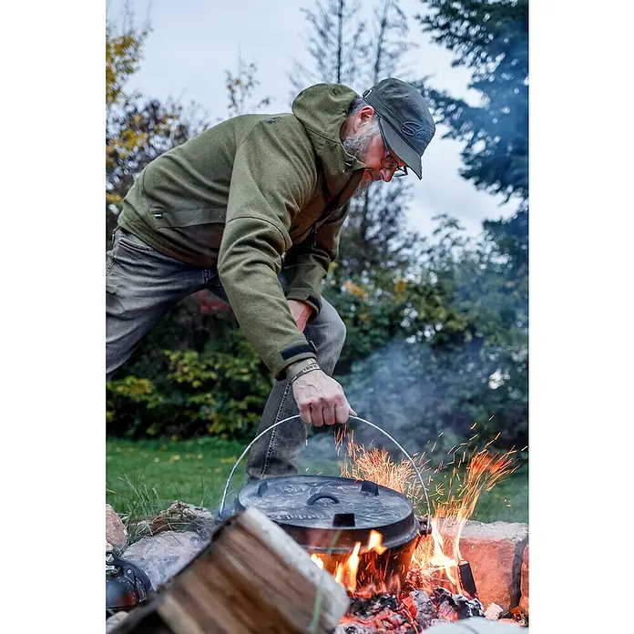 Petromax Dutch Oven - Ø x H: 42 x 20 cm, Gusseisen, 10,8 l, Planer Boden - Ein Mann mit Bart beugt sich über einen Dutch Oven über einem Lagerfeuer im Freien, umgeben von Bäumen und Grün.