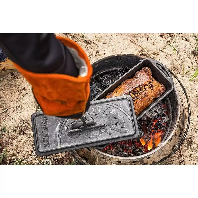 Petromax Auflaufform - L x B x H: 34,5 x 13 x 13,5 cm, Gusseisen, 2,4 l - Gusseiserne Brotbackform backt Brot in einer tragbaren Feuerschale. Eine Hand mit orangefarbenem Handschuh hält den verzierten Deckel der Form.
