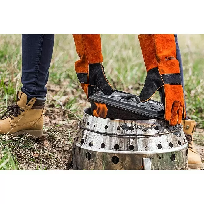Petromax Auflaufform - L x B x H: 34,5 x 13 x 13,5 cm, Gusseisen, 2,4 l - Person mit orangefarbenen Hitzeschutzhandschuhen platziert eine schwarze Gusseisenpfanne auf einem heißen silbernen Outdoor-Kocher mit Flammen.