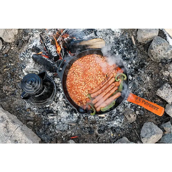 Petromax Handschutzgriff - Passend für: Petromax Grillpfannen, Leder/Aramidfasern - Lagerfeuer-Mahlzeit mit Petromax-Pfanne: Bohnen, Würstchen und Paprika brutzeln über dem offenen Feuer, während ein schwarzer Wasserkessel daneben steht.