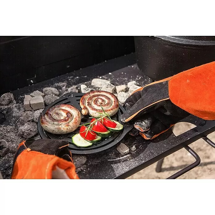 Petromax Grillrost mit Füßen - Gusseisen, Ø x H: 23 x 4,8 cm, Schwarz - Schwarzer Gusseisen-Grilleinsatz mit Würstchen und Gemüse, von einer behandschuhten Hand über Kohlen zubereitet.