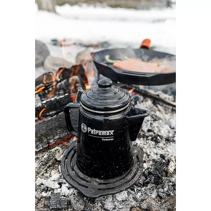 Petromax Grillrost mit Füßen - Gusseisen, Ø x H: 23 x 4,8 cm, Schwarz - Schwarze Petromax Perkomax Kaffeekanne auf einem Rost über Lagerfeuerkohlen, mit einer Bratpfanne in der Nähe.