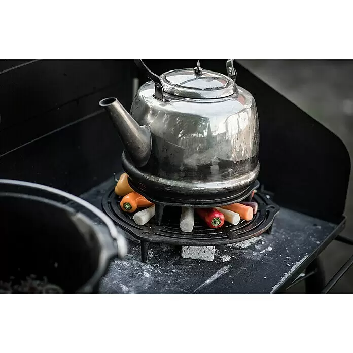 Petromax Grillrost mit Füßen - Gusseisen, Ø x H: 23 x 4,8 cm, Schwarz - Silberner Kessel auf Gusseisen-Untersetzer, wärmt bunte Paprika und Karotten auf einem Outdoor-Herd.