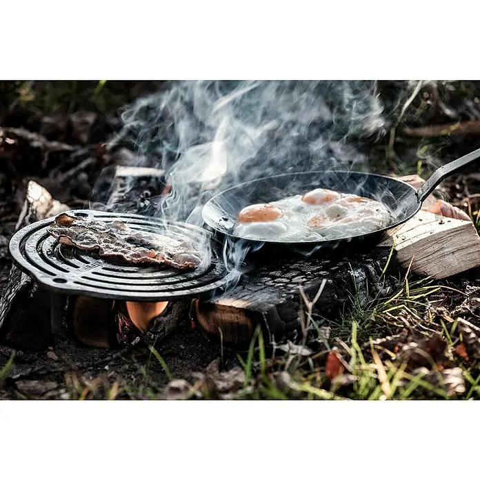 Petromax Grillrost mit Füßen - Gusseisen, Ø x H: 23 x 4,8 cm, Schwarz - Bacon und Spiegeleier werden im Freien auf einem Lagerfeuer zubereitet, was ein rustikales und köstliches Frühstückserlebnis bietet.