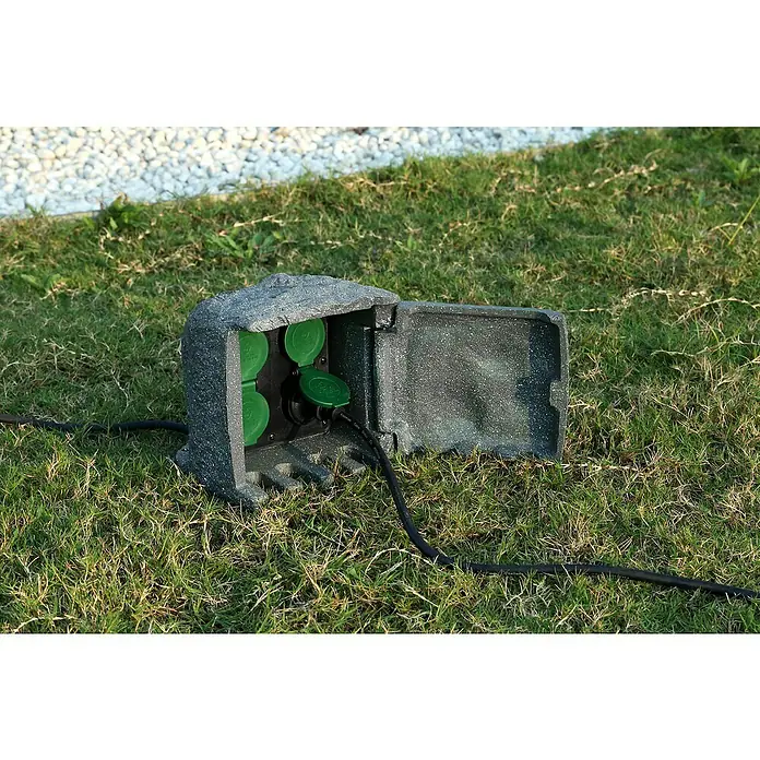 Voltomat Distribuidor de corriente para obras Power Rock - Número de enchufes Schuko: 4 ud., IP44 - Primer plano de una toma de corriente exterior resistente a la intemperie con aspecto de roca con tapa abierta, integrada en la hierba verde.