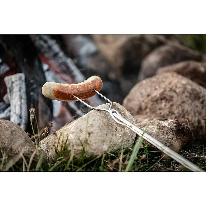 Petromax Grillspieß - Länge: 92 cm, Edelstahl/Buchenholz, Form: Gerade, 2 Stk. - Das Bild zeigt eine Wurst auf einem Grillspieß, die über einem Lagerfeuer gegrillt wird, umgeben von Steinen und Gras.