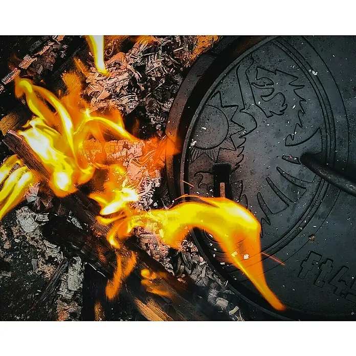 Petromax Dutch Oven - Ø x H: 31 x 14,6 cm, Gusseisen, 3,5 l, Planer Boden - Ein Dutch Oven Deckel mit einem Wolf- und Sonnenmuster liegt neben einem Lagerfeuer, das zum Kochen im Freien verwendet wird.