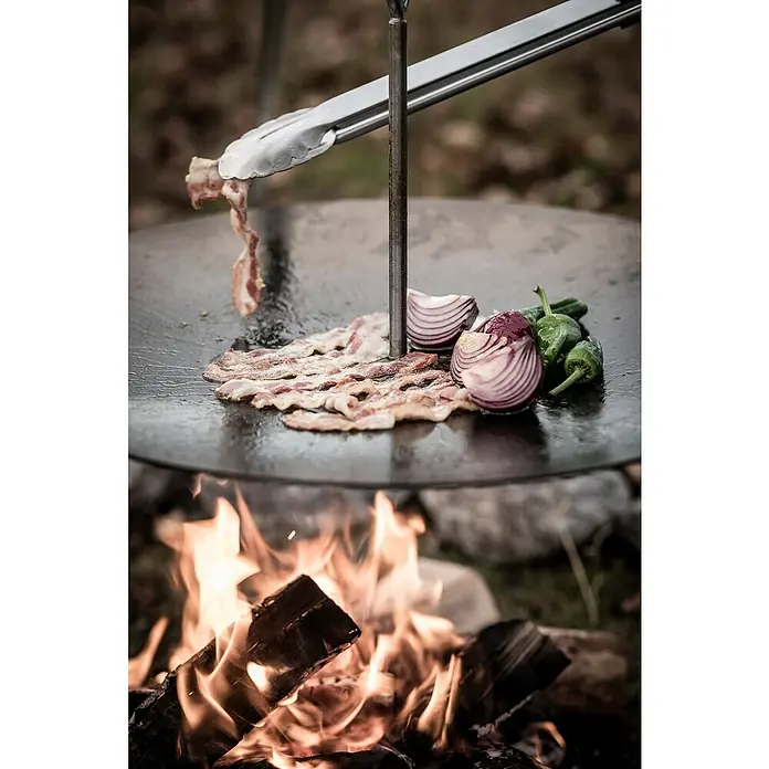 Petromax Grillzange - Länge: 54 cm - Grillplatte über offenem Feuer mit Speck, Zwiebeln und Paprika. Eine Grillzange hält ein Stück Speck über der Platte.