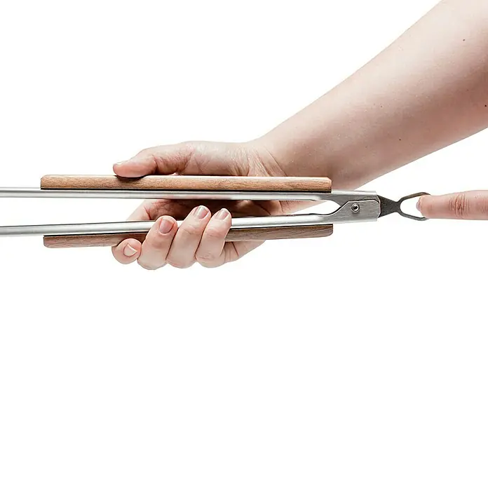Petromax Grillzange - Länge: 54 cm - Aufnahme einer Hand, die eine Grillzange mit Holzgriffen und Metallspitzen hält, vor einem schwarzen Hintergrund.