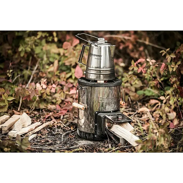 Petromax Holzkohlegrill Raketenofen - Grillfläche (B x T): 27,5 x 47,5 cm, Silber - Petromax Holzvergaserofen mit Edelstahl-Kaffeekanne auf dem Ofen, umgeben von herbstlichem Laub, ideal für Outdoor-Abenteuer und Kaffeegenuss.