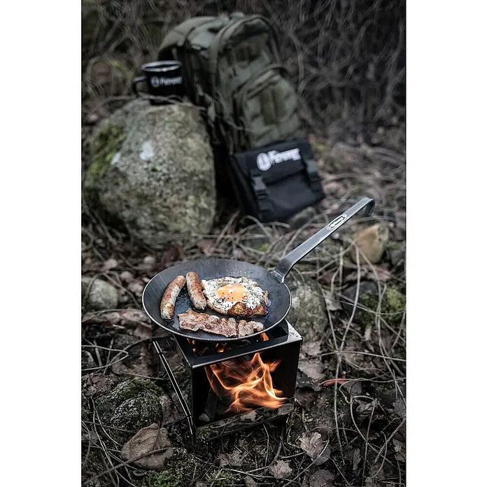 Petromax Grillpfanne - Durchmesser: 14 cm - 20 cm, Höhe: 12 cm, Eisen - Pfannengericht im Freien: Eine Bratpfanne auf einem Kocher mit Würstchen, Speck und Spiegelei, im Hintergrund Rucksack und Tasse.